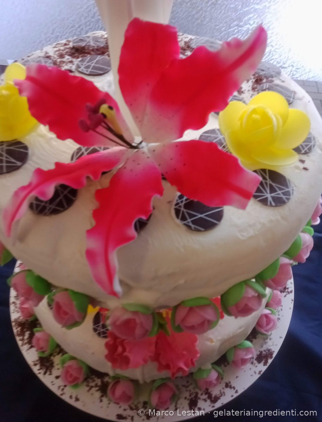Giglio in pasta di zucchero rosa su torta gelato bianca con rose di ostia gialle e dischi di cioccolato