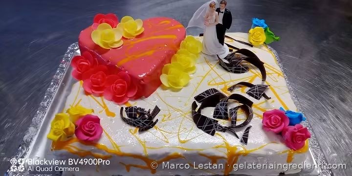 Torta rettangolare con glassa bianca, cuore rosa, rose gialle e rosse, scaglie di cioccolato e coppia di sposi